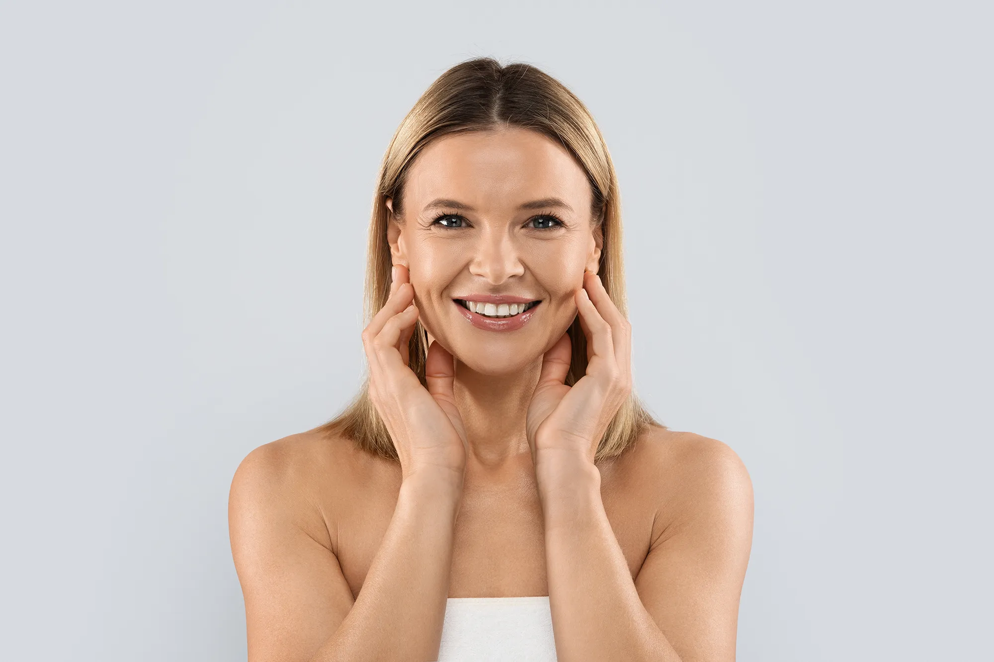 Femme souriante au teint lumineux touchant délicatement son visage sur fond gris clair dans un contexte esthétique et anti-âge