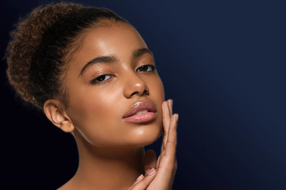 Femme au teint lumineux posant la main sur sa joue, sur fond bleu foncé.